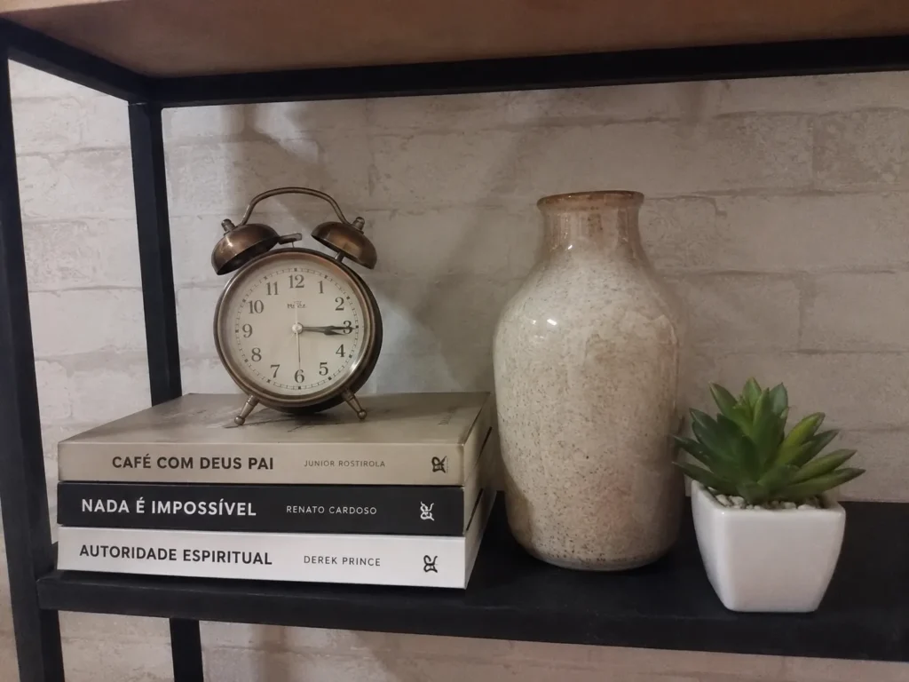 Detalhe de prateleira industrial preta com livros empilhados, relógio decorativo e vaso, demonstrando equilíbrio visual.
